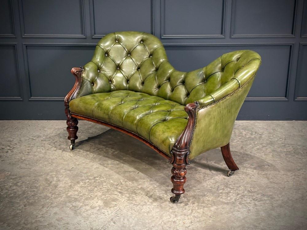 A Victorian double spoon-back settee upholstered in deep buttoned green leather with a carved mahogany frame set on brass castors. View VIII
