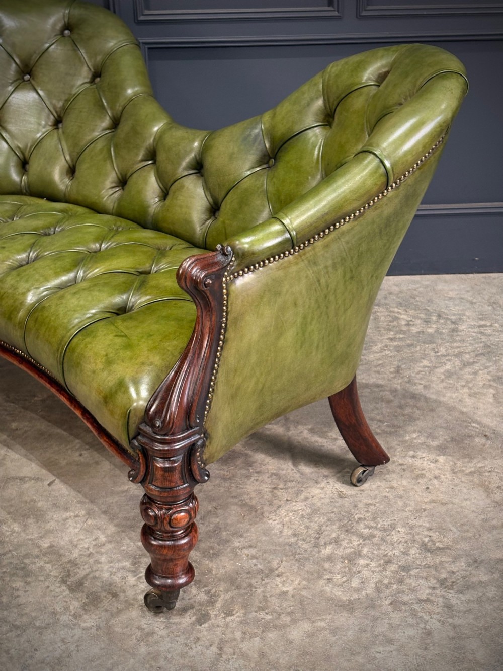 A Victorian double spoon-back settee upholstered in deep buttoned green leather with a carved mahogany frame set on brass castors. View IX