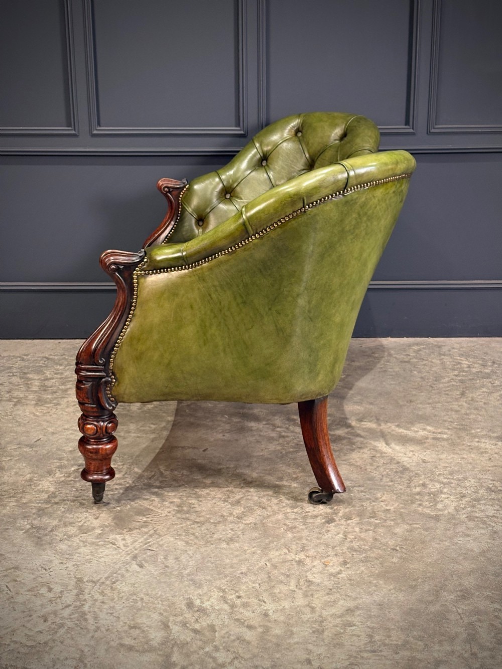 A Victorian double spoon-back settee upholstered in deep buttoned green leather with a carved mahogany frame set on brass castors. View X