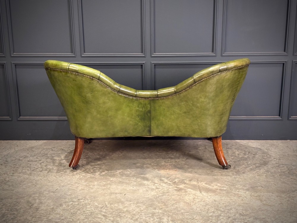A Victorian double spoon-back settee upholstered in deep buttoned green leather with a carved mahogany frame set on brass castors. View XI