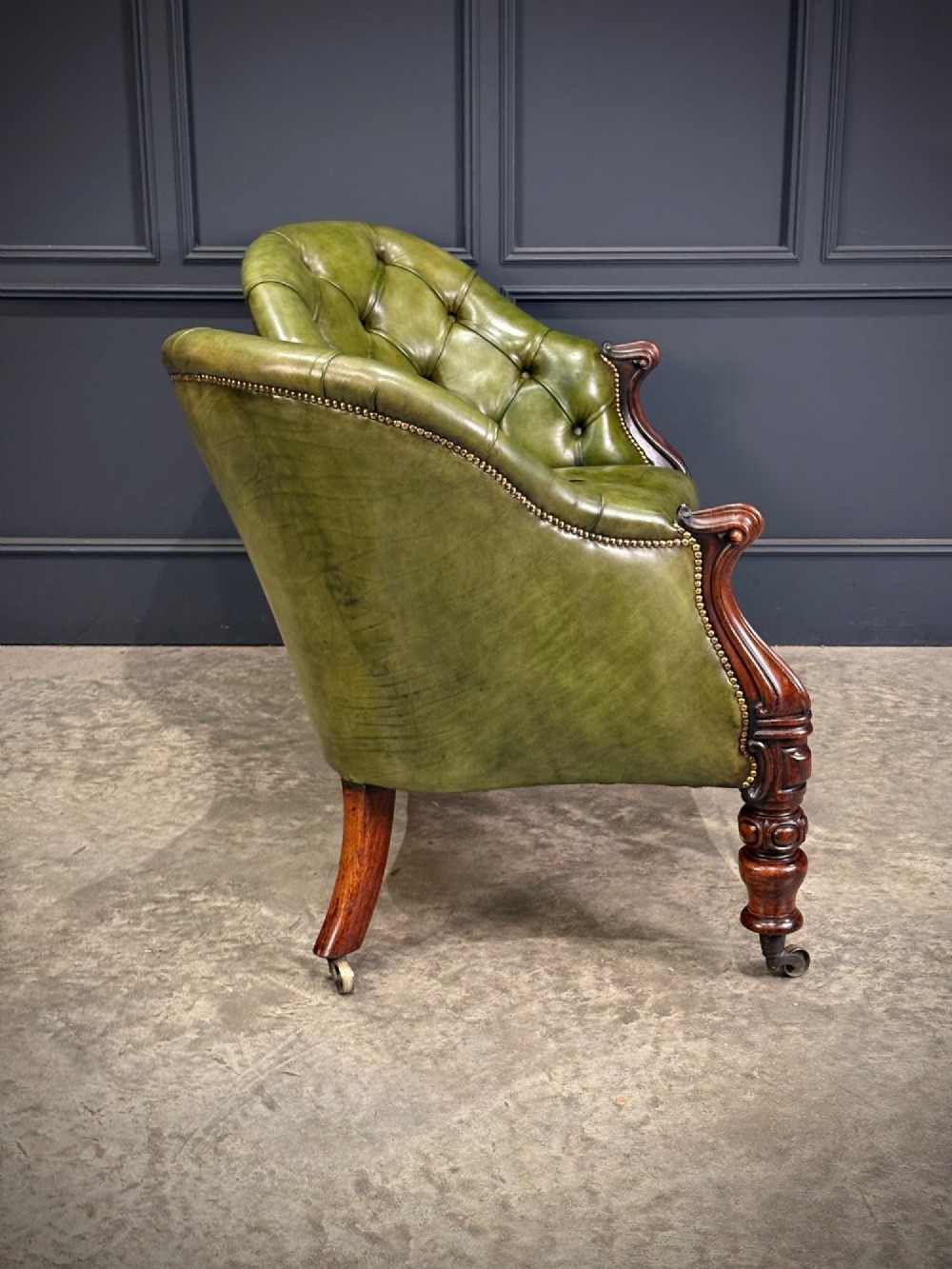 A Victorian double spoon-back settee upholstered in deep buttoned green leather with a carved mahogany frame set on brass castors. View XII