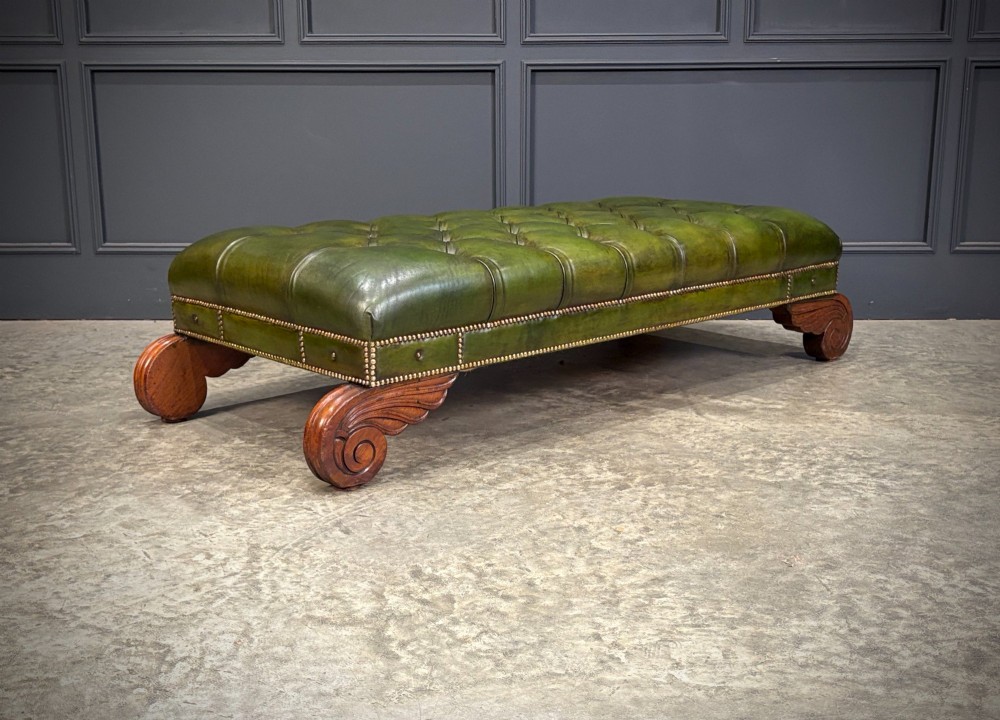 A low rectangular Regency style footstool upholstered in deep-buttoned green leather with brass stud detailing and carved wooden scroll feet. View II