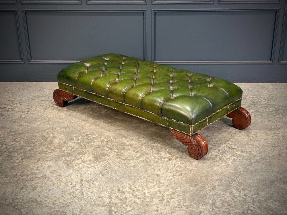 A low rectangular Regency style footstool upholstered in deep-buttoned green leather with brass stud detailing and carved wooden scroll feet. View VI
