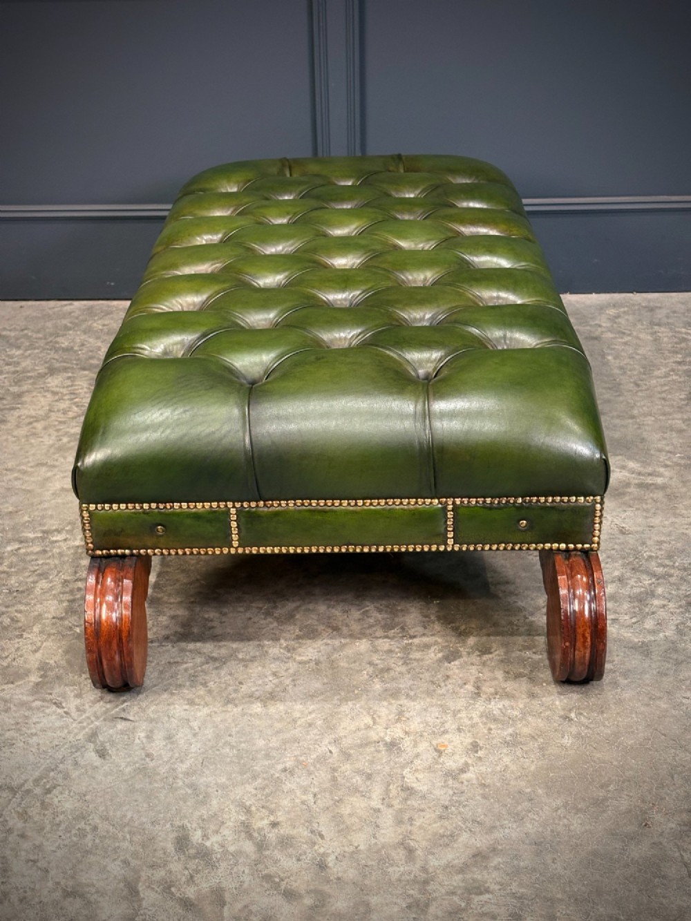 A low rectangular Regency style footstool upholstered in deep-buttoned green leather with brass stud detailing and carved wooden scroll feet. View VII