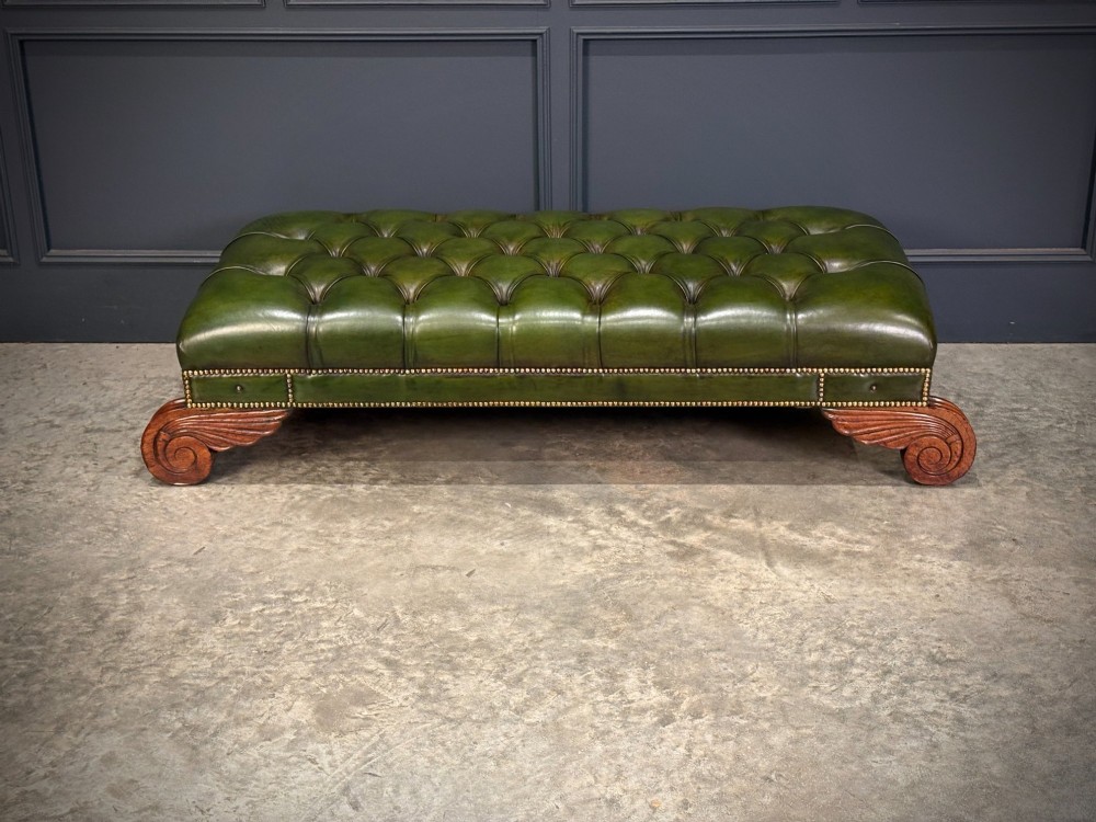 A low rectangular Regency style footstool upholstered in deep-buttoned green leather with brass stud detailing and carved wooden scroll feet. View IX