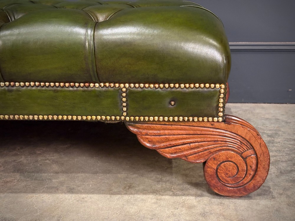 A low rectangular Regency style footstool upholstered in deep-buttoned green leather with brass stud detailing and carved wooden scroll feet. View X