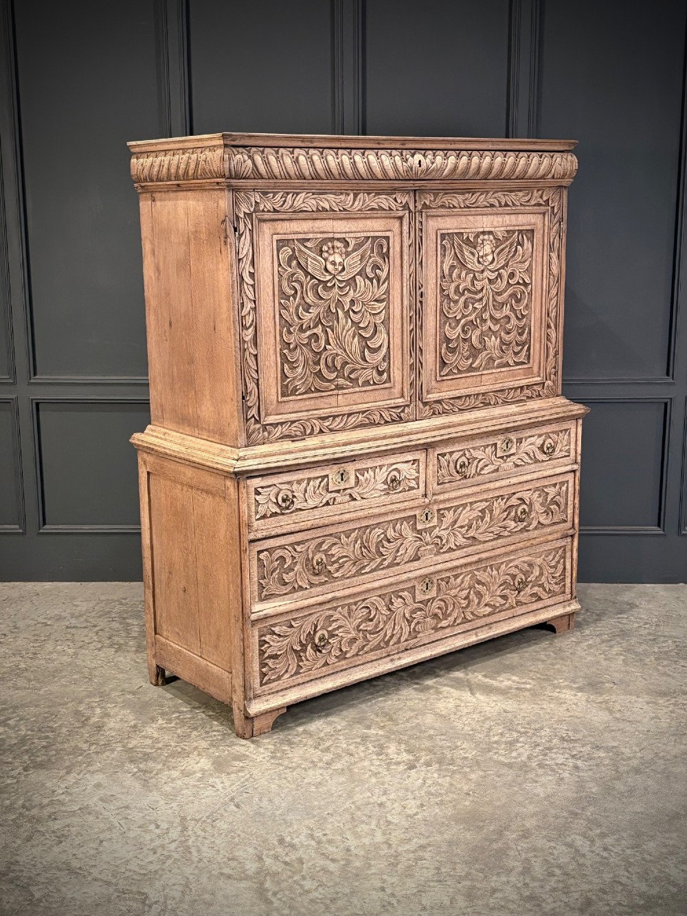 An 18th-century light oak two-piece cupboard on chest featuring detailed hand-carved foliate panels, cherub motifs, and carved drawer fronts.