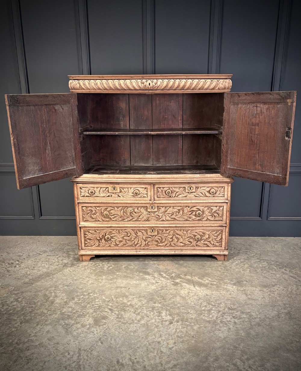 An 18th-century light oak two-piece cupboard on chest featuring detailed hand-carved foliate panels, cherub motifs, and carved drawer fronts. View V
