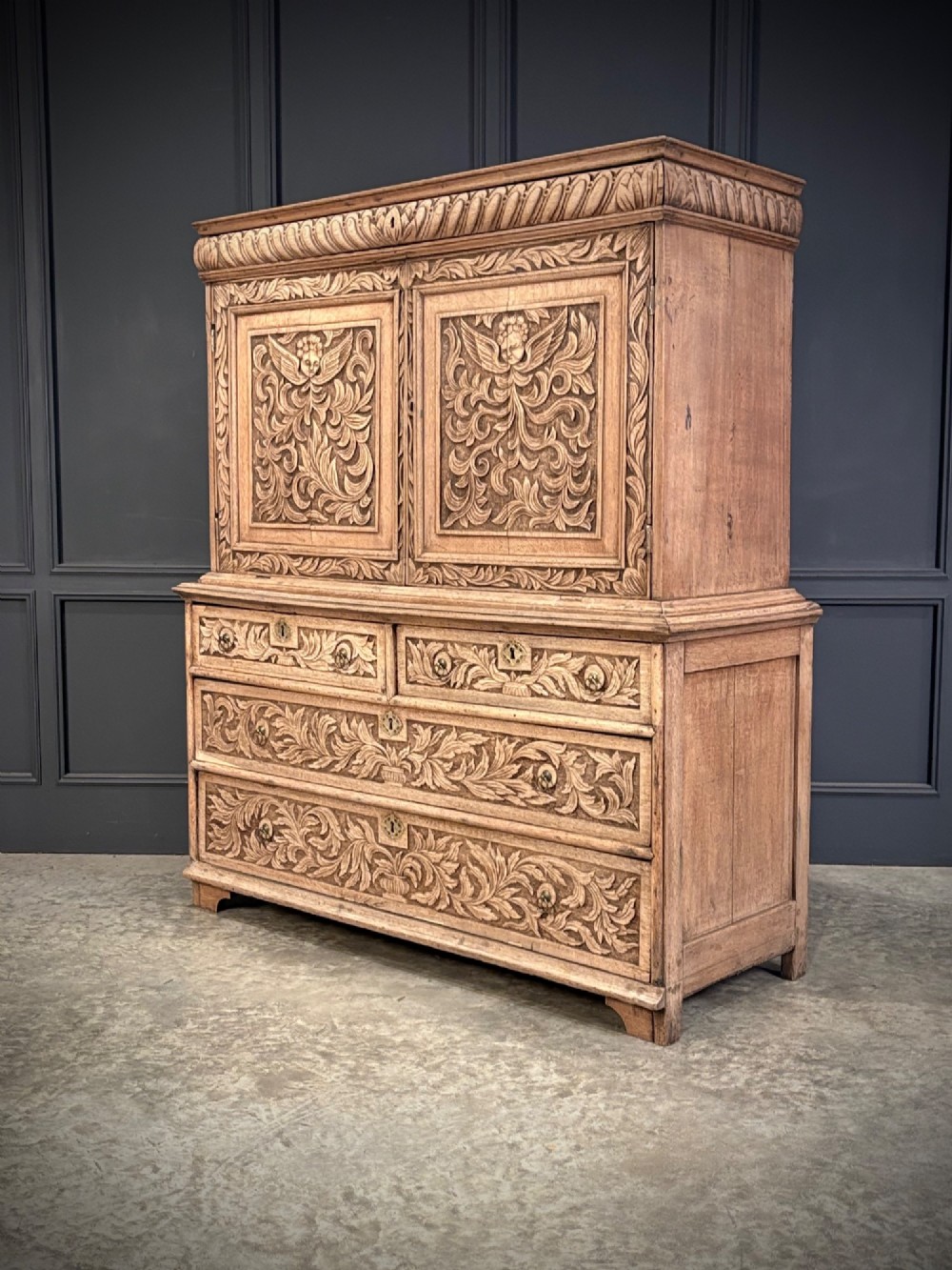 An 18th-century light oak two-piece cupboard on chest featuring detailed hand-carved foliate panels, cherub motifs, and carved drawer fronts. View VI