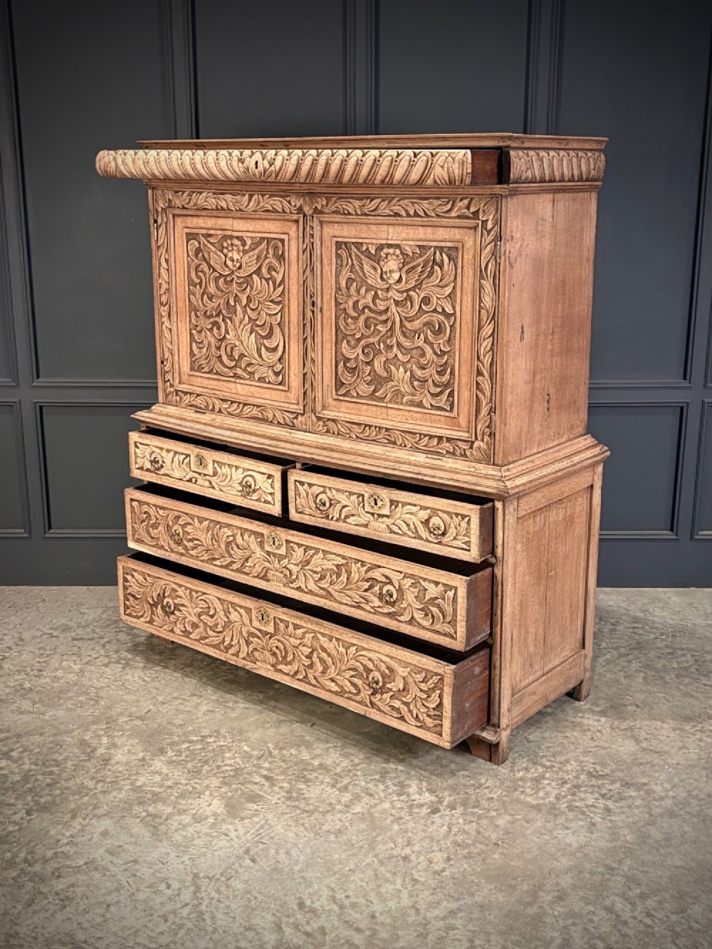 An 18th-century light oak two-piece cupboard on chest featuring detailed hand-carved foliate panels, cherub motifs, and carved drawer fronts. View VII