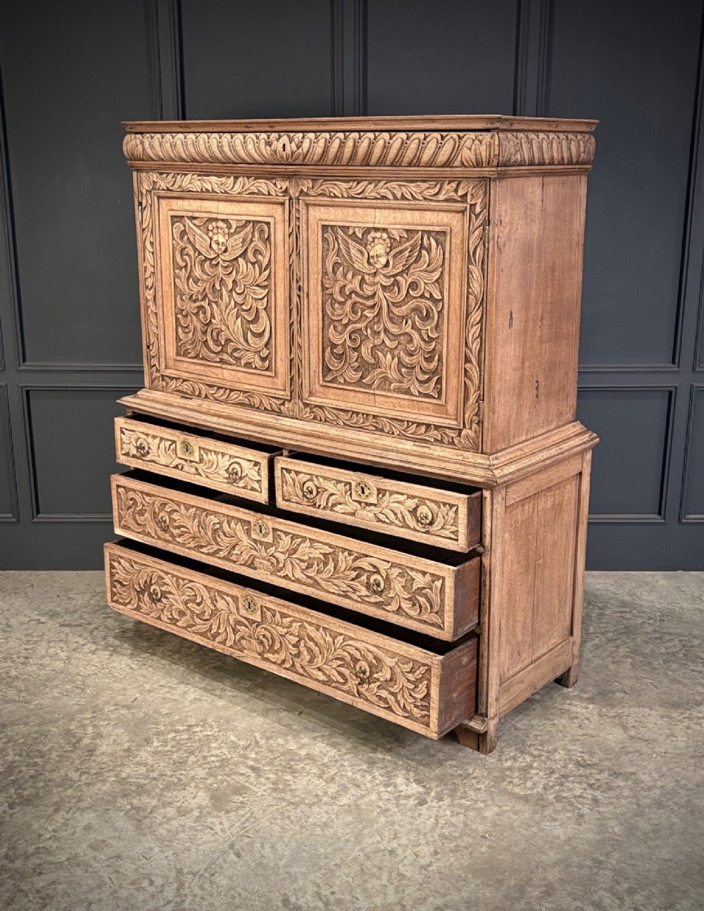An 18th-century light oak two-piece cupboard on chest featuring detailed hand-carved foliate panels, cherub motifs, and carved drawer fronts. View VIII