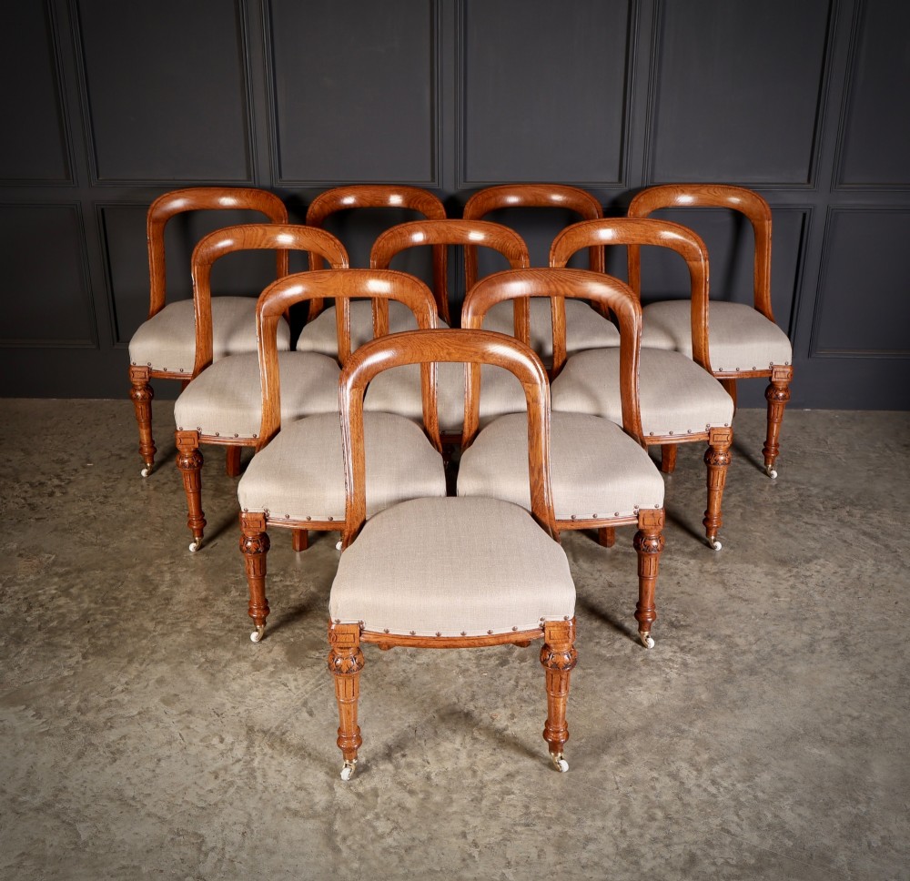 A set of ten Victorian oak dining chairs with rounded hoop backs, neutral fabric seats with stud trim, and carved front legs on ceramic castors.