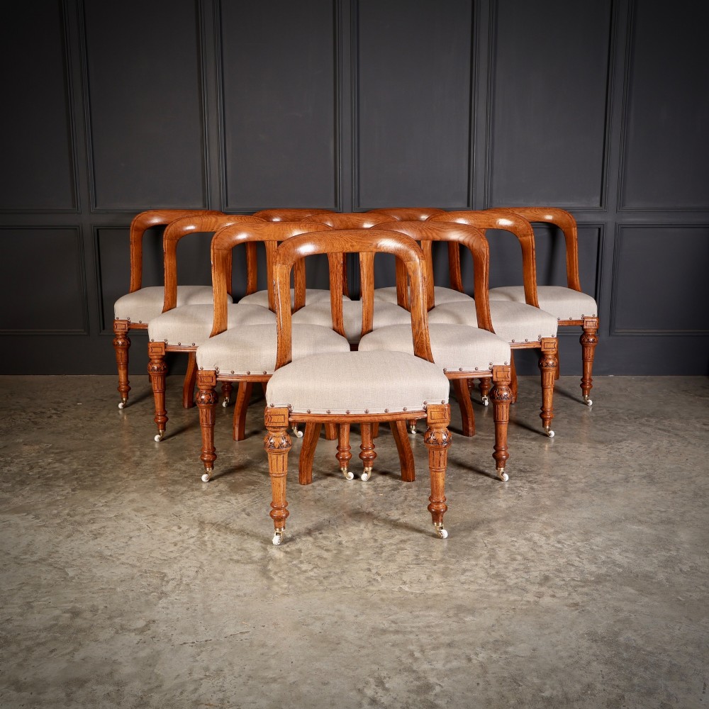 A set of ten Victorian oak dining chairs with rounded hoop backs, neutral fabric seats with stud trim, and carved front legs on ceramic castors. View II