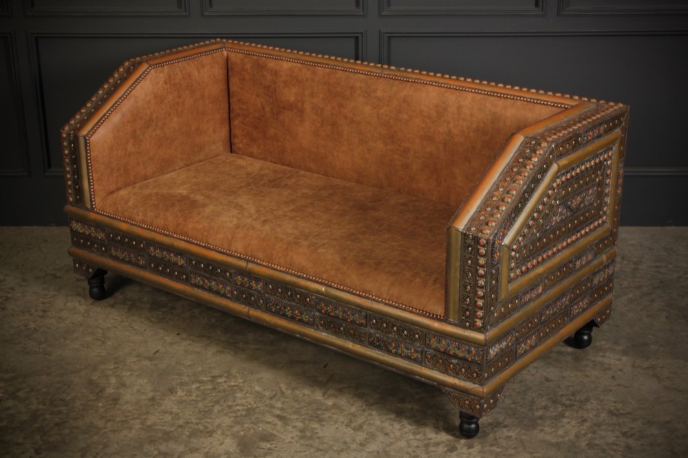 A boxy Moroccan-style sofa featuring tan leather upholstery on the seat and interior, surrounded by an exterior clad in embossed brass metal panels with decorative studding. View VI