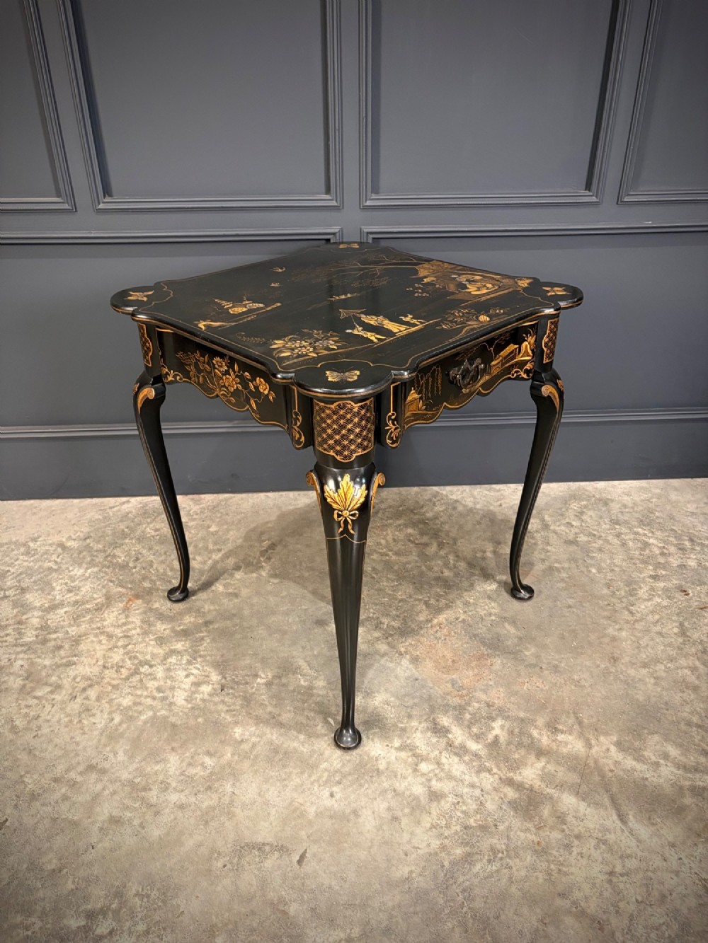 A black lacquered square occasional table featuring gilded Chinoiserie landscape scenes, shaped apron edges, a single drawer, and slender cabriole legs.