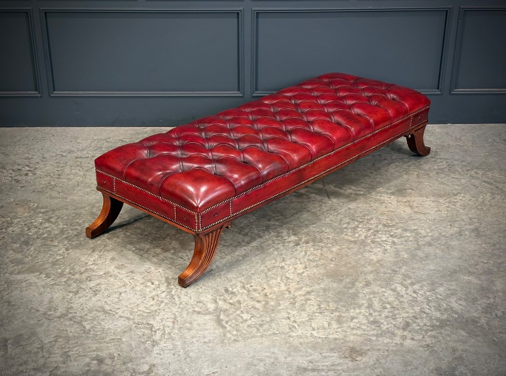 A long rectangular footstool upholstered in deep-buttoned red leather with brass studding along the trim and carved hardwood legs.