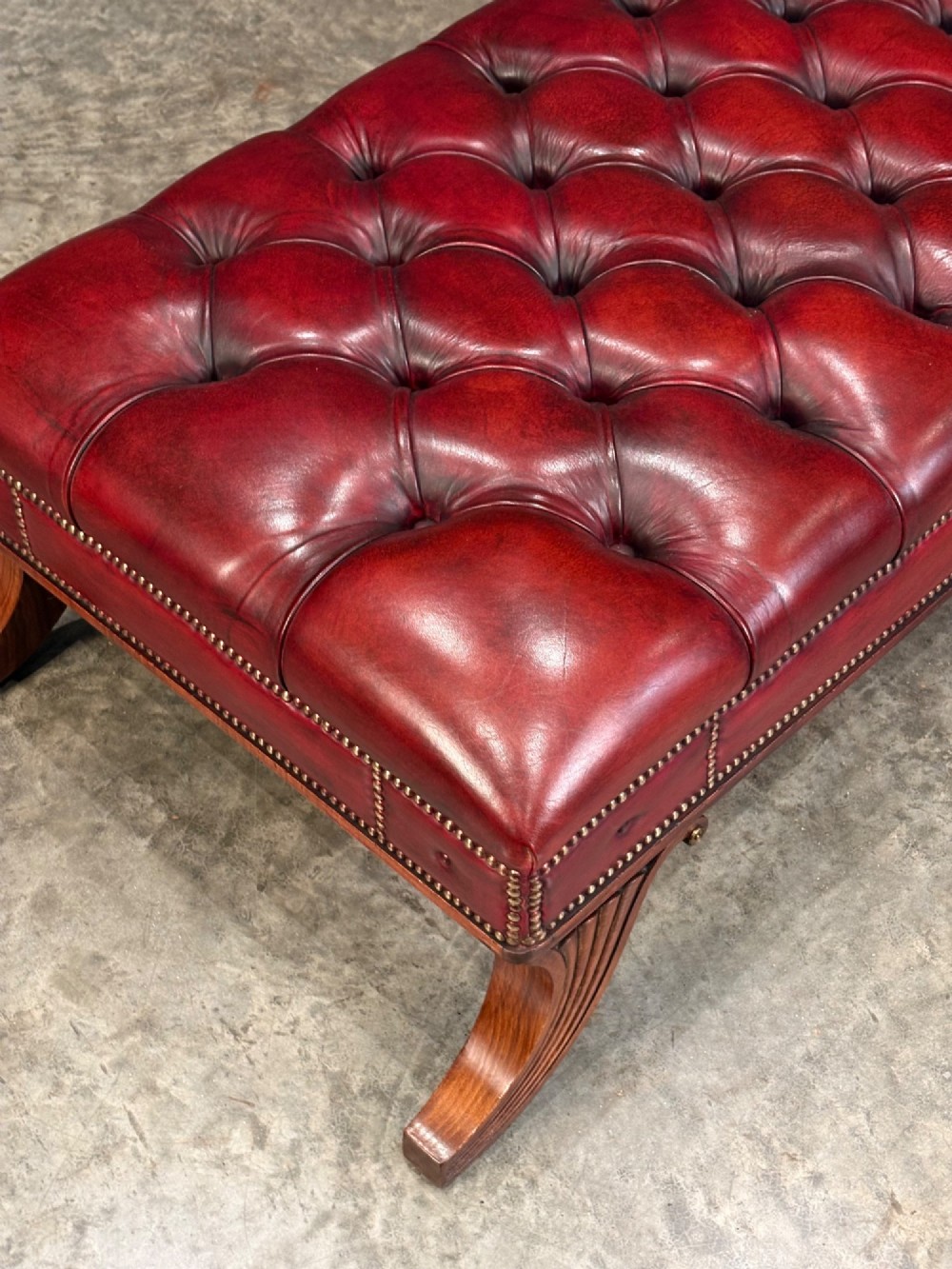 A long rectangular footstool upholstered in deep-buttoned red leather with brass studding along the trim and carved hardwood legs. View II