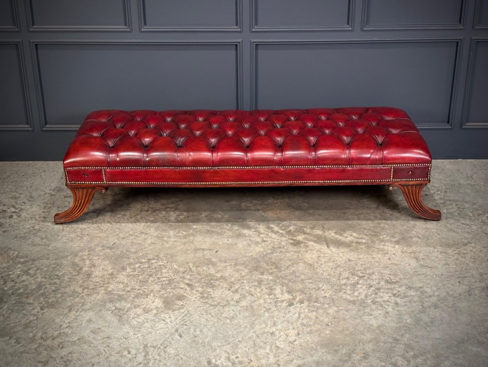 A long rectangular footstool upholstered in deep-buttoned red leather with brass studding along the trim and carved hardwood legs. View III