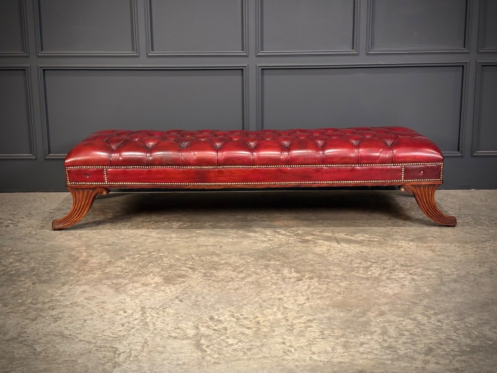 A long rectangular footstool upholstered in deep-buttoned red leather with brass studding along the trim and carved hardwood legs. View IV