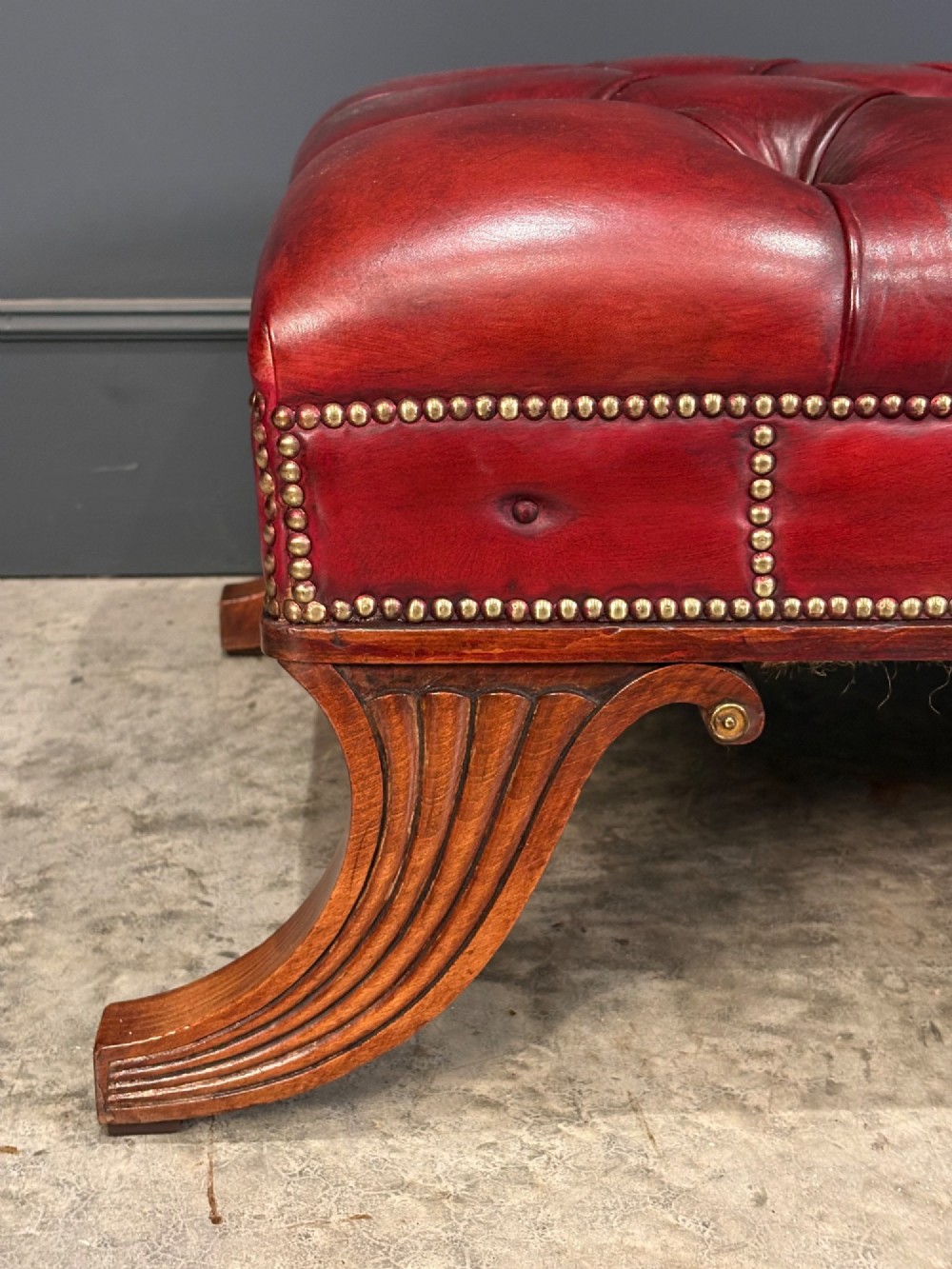 A long rectangular footstool upholstered in deep-buttoned red leather with brass studding along the trim and carved hardwood legs. View V