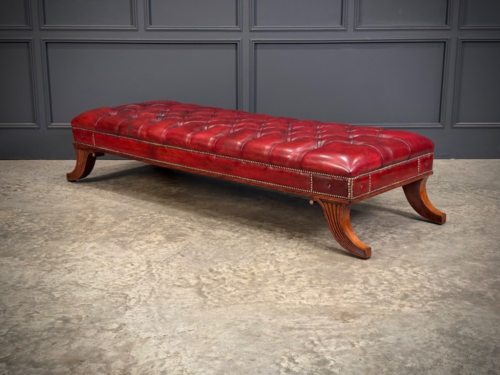 A long rectangular footstool upholstered in deep-buttoned red leather with brass studding along the trim and carved hardwood legs. View VI