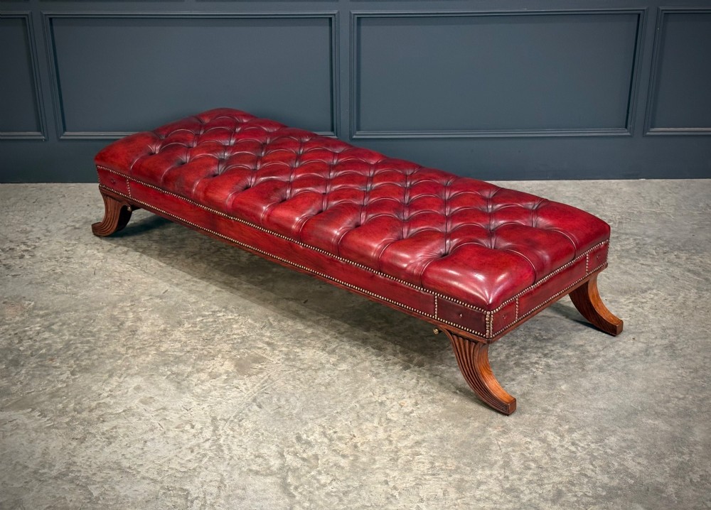 A long rectangular footstool upholstered in deep-buttoned red leather with brass studding along the trim and carved hardwood legs. View VII