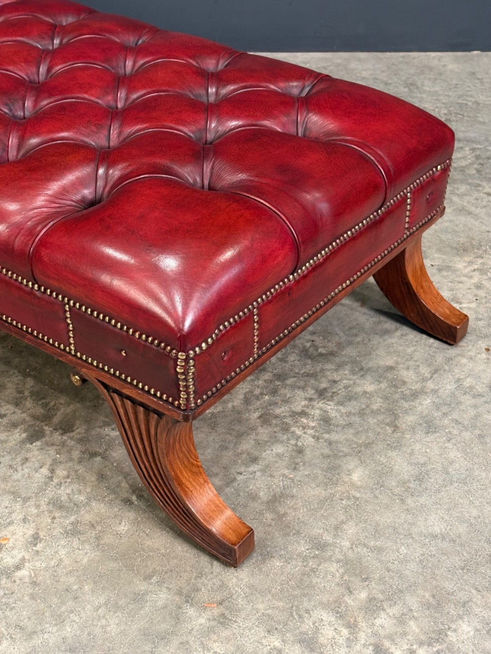A long rectangular footstool upholstered in deep-buttoned red leather with brass studding along the trim and carved hardwood legs. View VIII