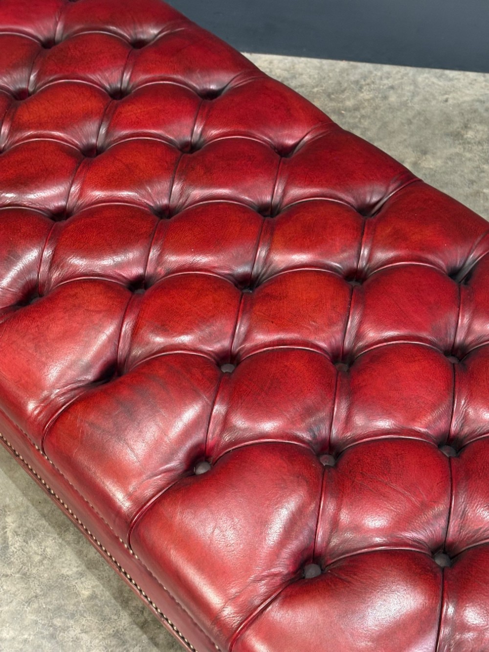 A long rectangular footstool upholstered in deep-buttoned red leather with brass studding along the trim and carved hardwood legs. View IX