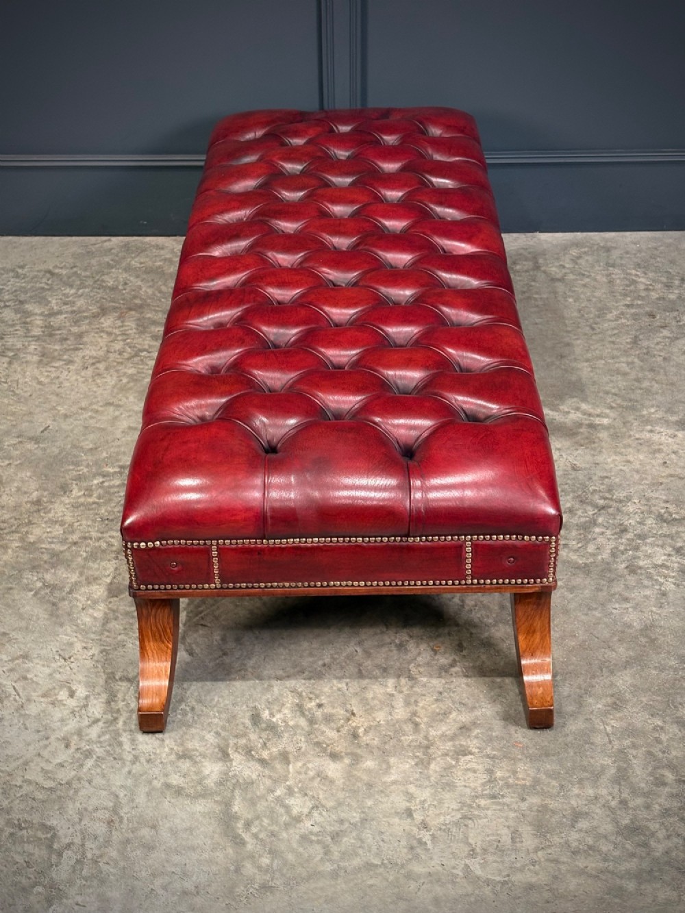 A long rectangular footstool upholstered in deep-buttoned red leather with brass studding along the trim and carved hardwood legs. View X