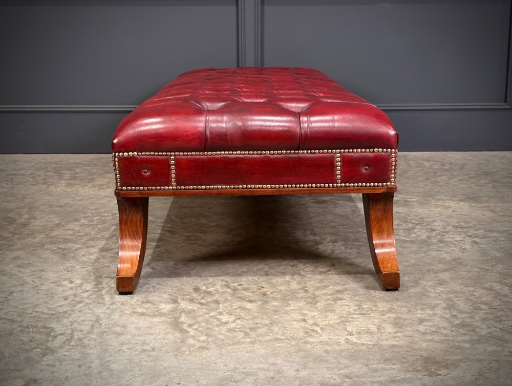 A long rectangular footstool upholstered in deep-buttoned red leather with brass studding along the trim and carved hardwood legs. View XI