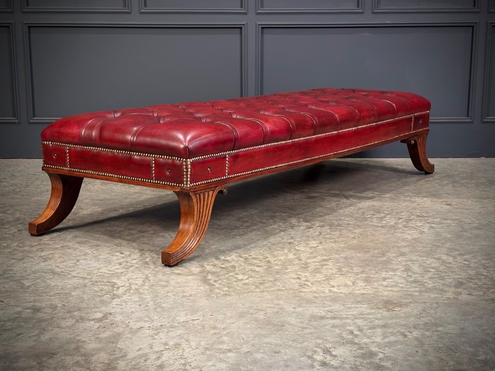 A long rectangular footstool upholstered in deep-buttoned red leather with brass studding along the trim and carved hardwood legs. View XII