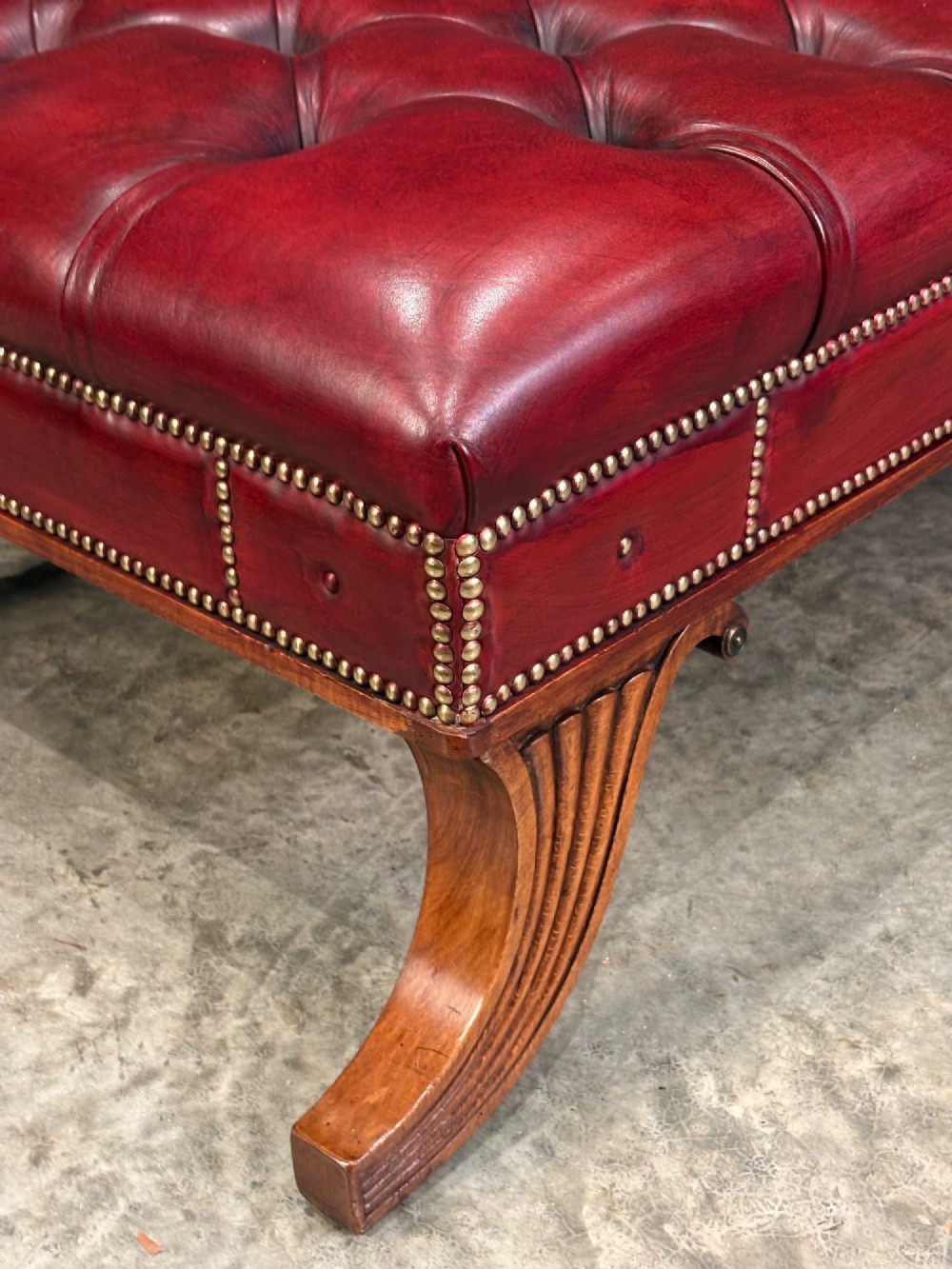 A long rectangular footstool upholstered in deep-buttoned red leather with brass studding along the trim and carved hardwood legs. View XIII