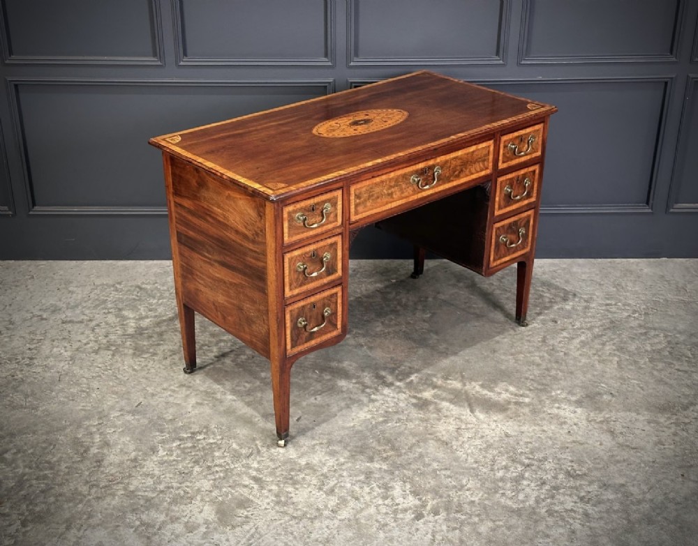 An eighteenth-century mahogany writing desk featuring a central oval floral marquetry inlay on top, crossbanded borders, seven drawers with brass handles, and square tapered legs.
