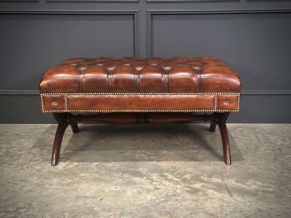 A rectangular hand-dyed brown leather footstool with deep button tufting, brass studding, and an X-frame wooden base. View II