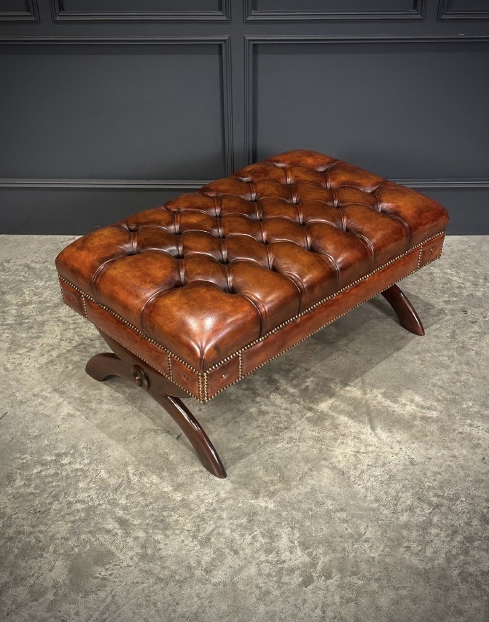 A rectangular hand-dyed brown leather footstool with deep button tufting, brass studding, and an X-frame wooden base.