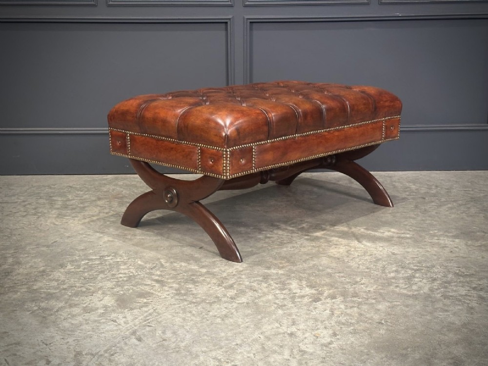A rectangular hand-dyed brown leather footstool with deep button tufting, brass studding, and an X-frame wooden base. View V