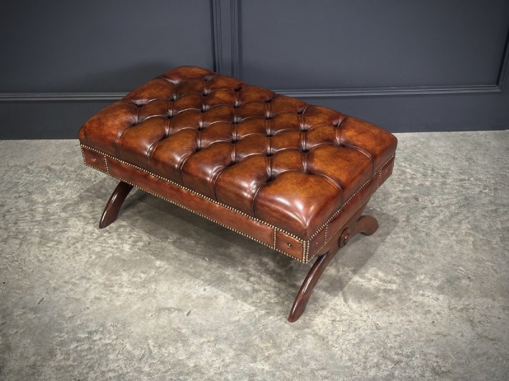 A rectangular hand-dyed brown leather footstool with deep button tufting, brass studding, and an X-frame wooden base. View VI