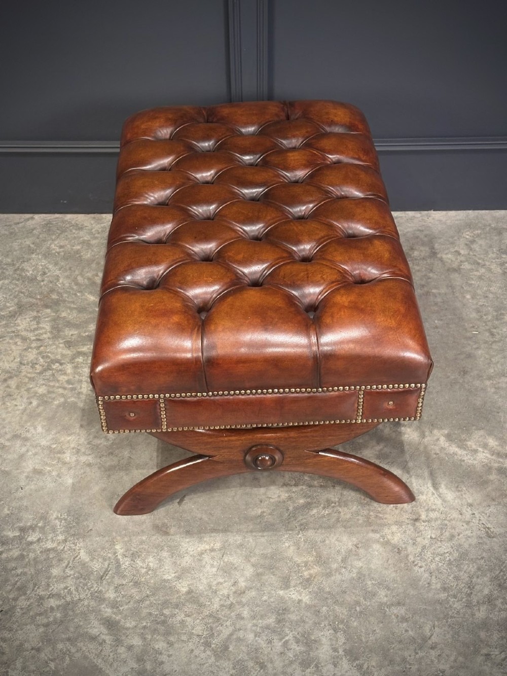 A rectangular hand-dyed brown leather footstool with deep button tufting, brass studding, and an X-frame wooden base. View VII