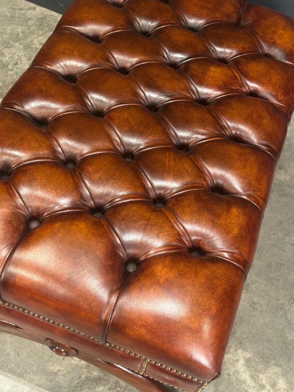 A rectangular hand-dyed brown leather footstool with deep button tufting, brass studding, and an X-frame wooden base. View VIII