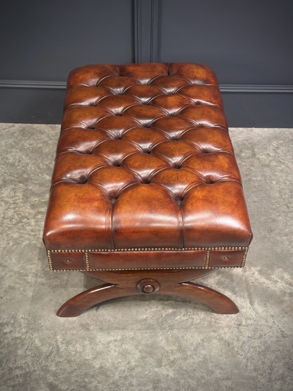 A rectangular hand-dyed brown leather footstool with deep button tufting, brass studding, and an X-frame wooden base. View X