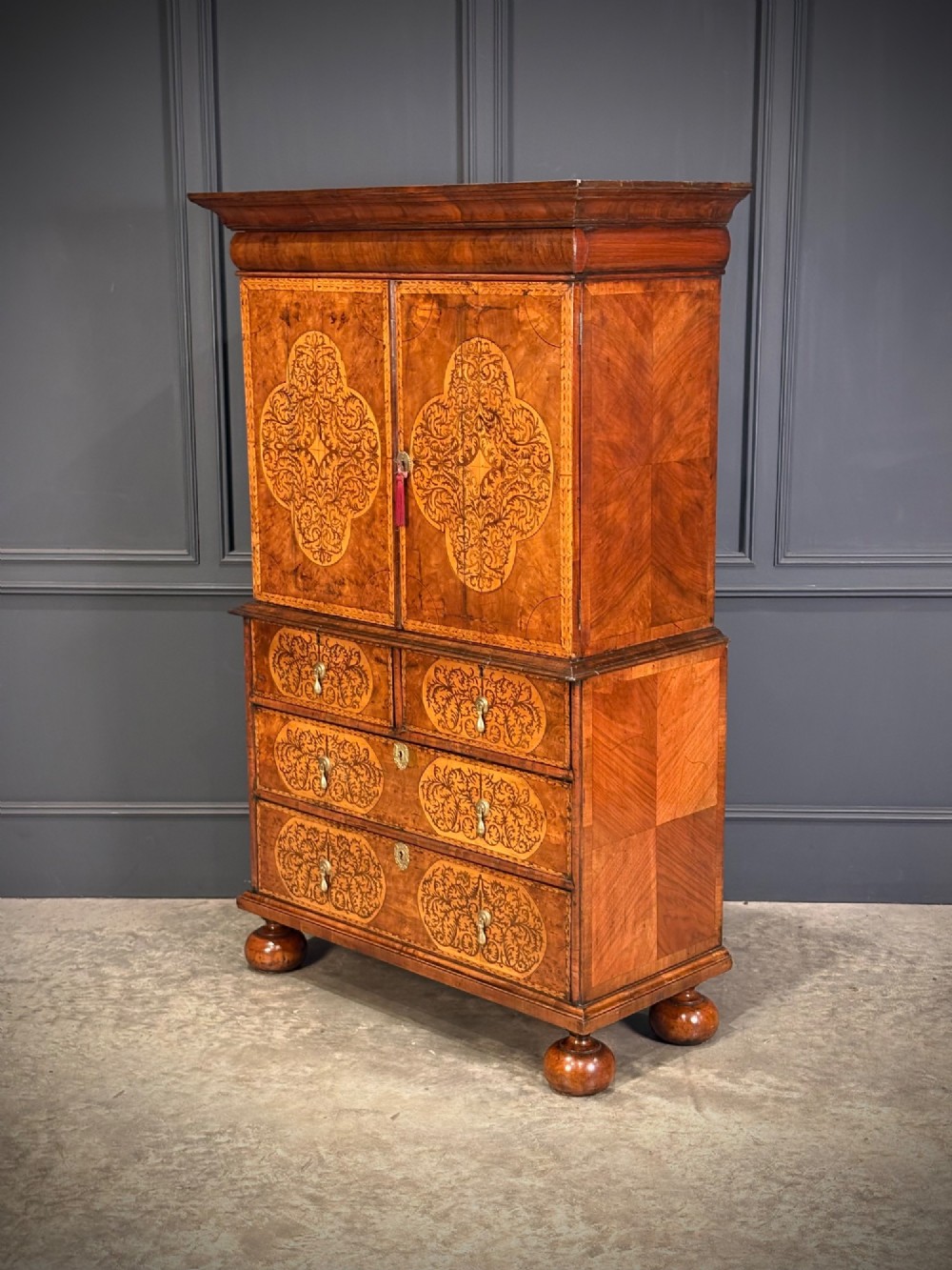A William and Mary period burr walnut cabinet on stand featuring detailed floral marquetry inlays, teardrop brass handles, and heavy bun feet. View VI