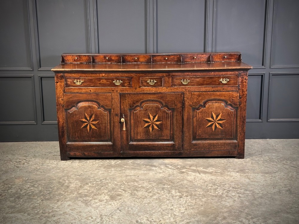 An 18th-century dark oak dresser base featuring three main drawers over three arched cupboard doors with star inlay details and brass handles.