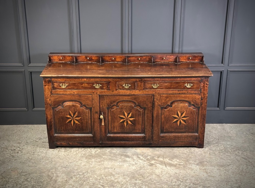 An 18th-century dark oak dresser base featuring three main drawers over three arched cupboard doors with star inlay details and brass handles. View IV