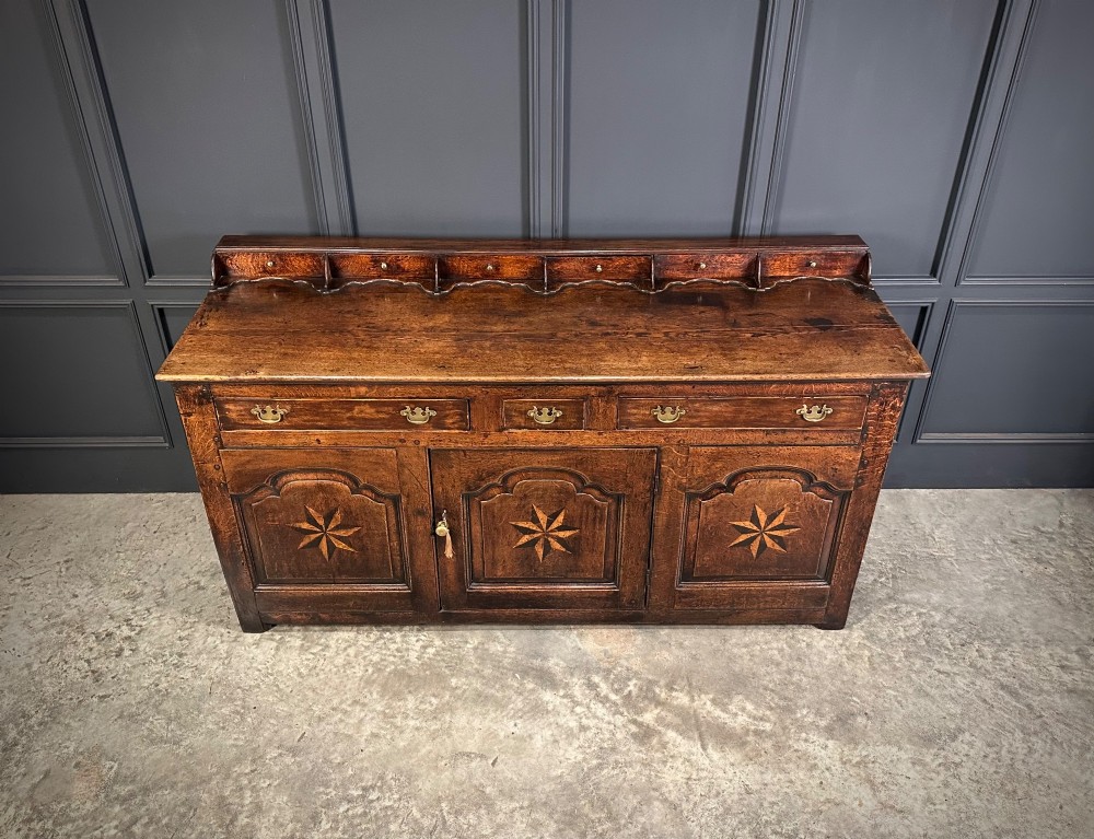 An 18th-century dark oak dresser base featuring three main drawers over three arched cupboard doors with star inlay details and brass handles. View V