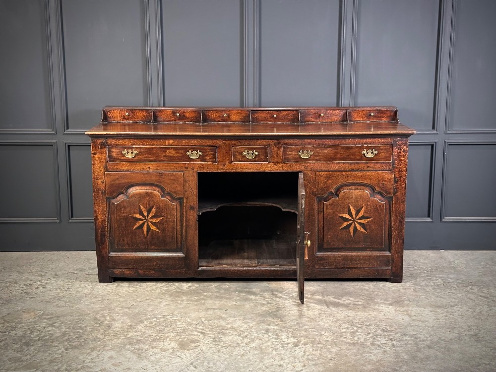 An 18th-century dark oak dresser base featuring three main drawers over three arched cupboard doors with star inlay details and brass handles. View VI