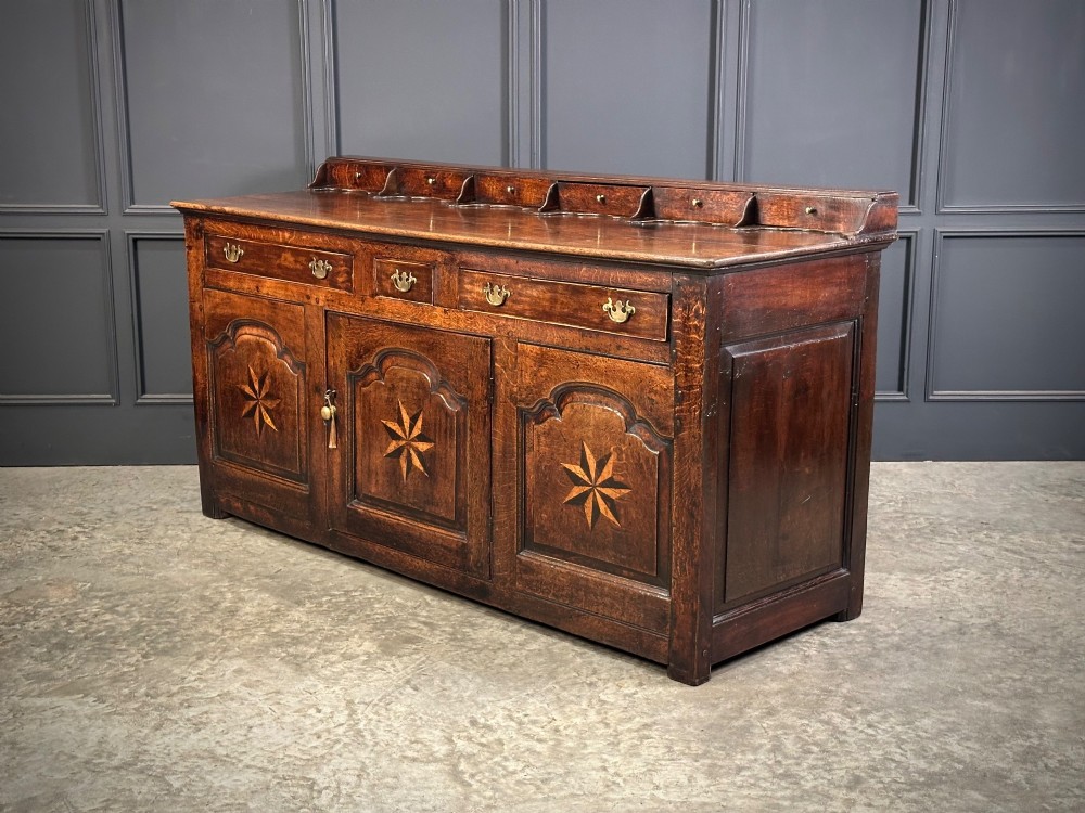 An 18th-century dark oak dresser base featuring three main drawers over three arched cupboard doors with star inlay details and brass handles. View VII