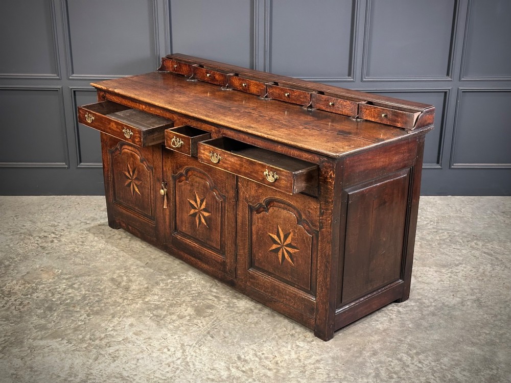 An 18th-century dark oak dresser base featuring three main drawers over three arched cupboard doors with star inlay details and brass handles. View VIII