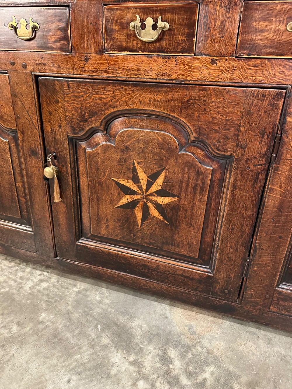 An 18th-century dark oak dresser base featuring three main drawers over three arched cupboard doors with star inlay details and brass handles. View XII