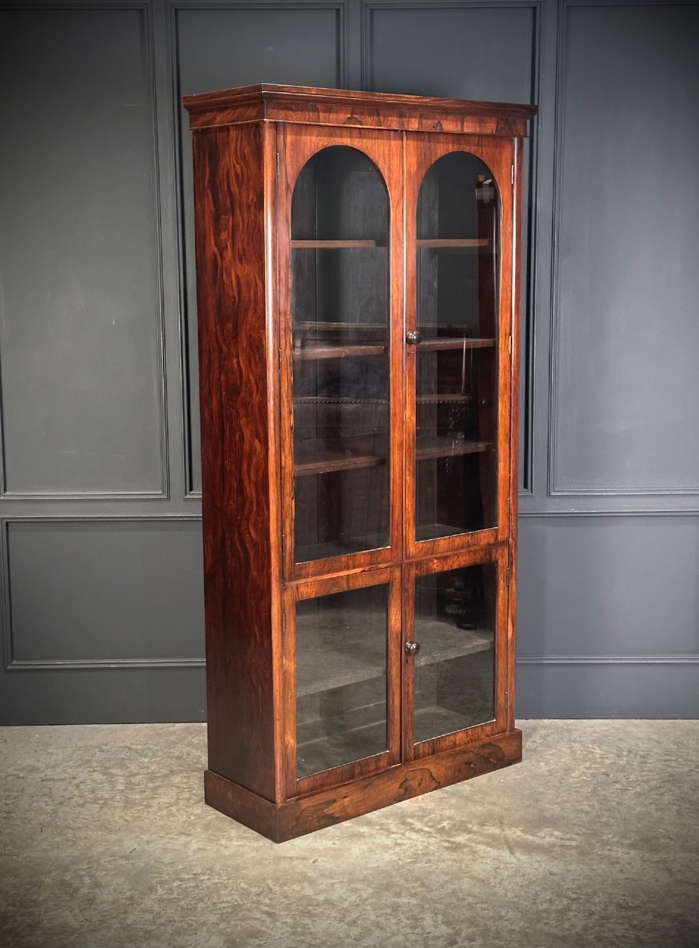 A tall Victorian rosewood bookcase featuring two upper arched glass doors and two lower glass doors on a flat plinth base.