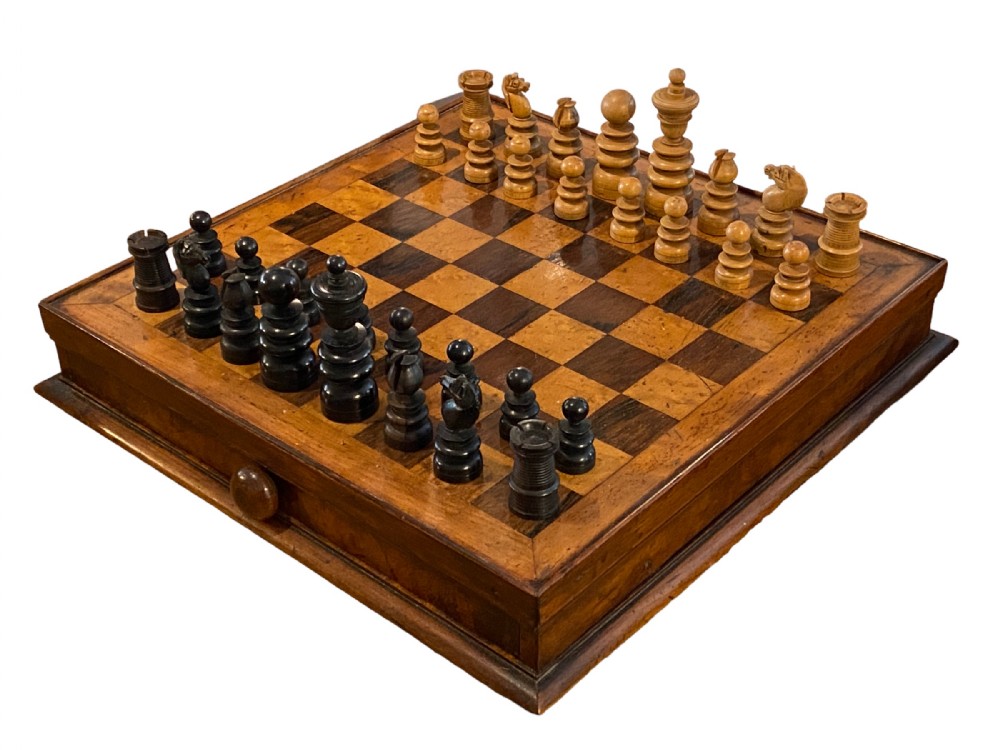 An antique nineteenth-century wooden chess compendium with an inlaid chessboard top, a storage drawer beneath, and carved light and dark wooden chess pieces arranged on the board.